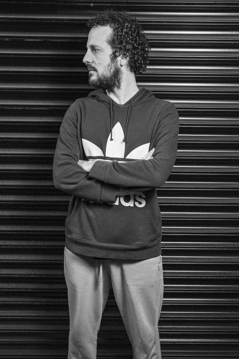 Retrato preto e branco do baterista Bruno Pigat, de braços cruzados e olhando para o lado, em frente ao portão texturizado. (Banda Madressilva, Ensaio Musical, Fotos Bruno Arita)'