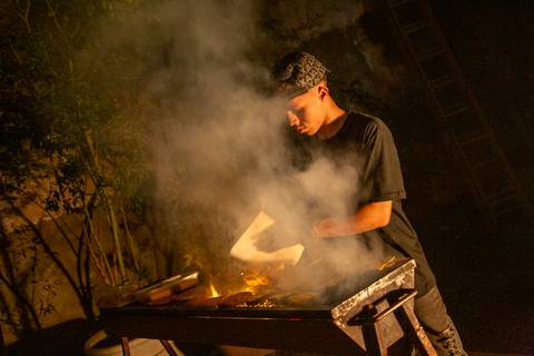 Churrasqueiro preparando hambúrgueres e espetinhos com fumaça, em foto artística que destaca a área de comidas do evento. (Ninfa Flores, São Paulo, Fotos Bruno Arita)'