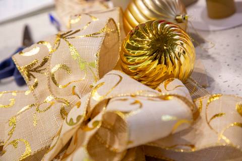 Plano detalhe de um laço branco com detalhes dourados e bolhas de Natal douradas ao lado, itens usados no workshop. (Westwing Moema, São Paulo, Fotos Bruno Arita)'