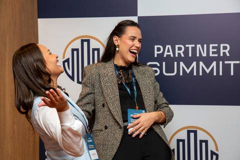Duas participantes rindo em momento espontâneo de networking em frente ao backdrop do Partner Summit. (Grand Hyatt SP, Evento Corporativo, Fotos Bruno Arita)'