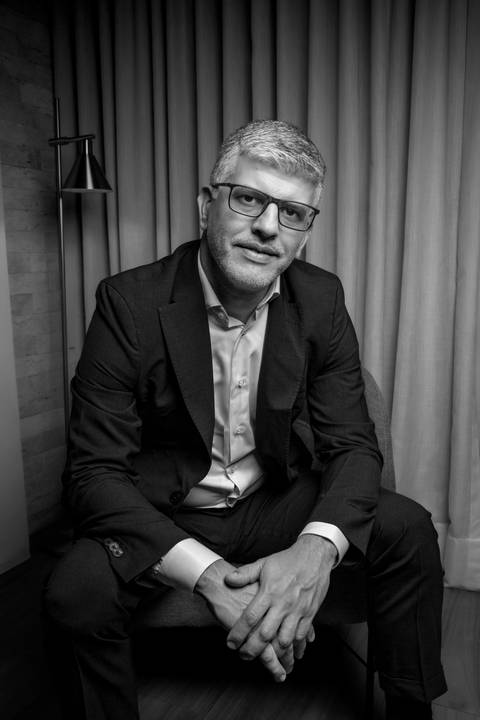 Retrato editorial em preto e branco de Fábio Rocha sentado em poltrona, mãos entrelaçadas, vestindo blazer e camisa em ambiente interno sofisticado com cortinas. (Branding Pessoal, Fotos Bruno Arita)'