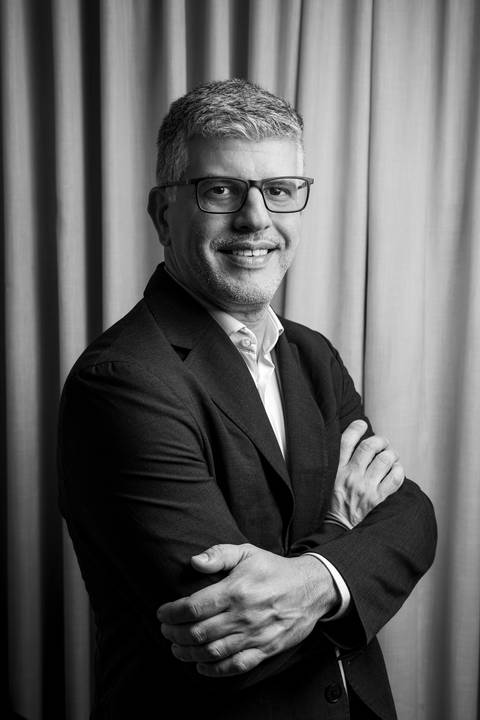 Fábio Rocha em retrato vertical P&B de braços cruzados e sorriso confiante, unindo autoridade e leveza em cenário de interior. (LinkedIn, Fotos Bruno Arita)'