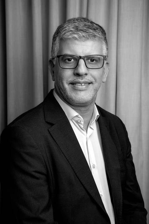 Close-up em preto e branco de Fábio Rocha com foco no rosto, vestindo blazer escuro, camisa clara e óculos, sorrindo amigavelmente com ar profissional. (LinkedIn, Fotos Bruno Arita)'