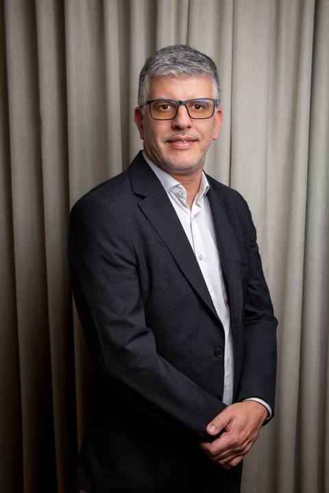 Plano médio vertical de Fábio Rocha com mãos postas à frente, vestindo traje social em cenário residencial autêntico com fundo neutro. (Ensaio Editorial, Fotos Bruno Arita)'