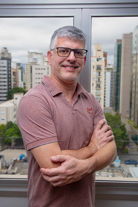 Fábio Rocha de braços cruzados e sorriso aberto, vestindo camisa polo bordô em frente a uma janela com vista para a cidade de São Paulo. (Lifestyle Profissional, Fotos Bruno Arita)'