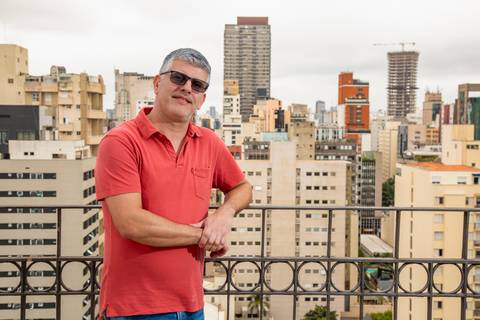 Fábio Rocha posando de forma descontraída em terraço paulistano, vestindo camisa polo coral e óculos, com o skyline da cidade ao fundo. (Retrato Editorial, Fotos Bruno Arita)'