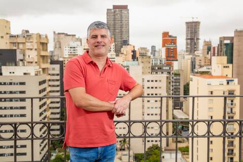 Fábio Rocha em plano médio horizontal, vestindo camisa polo coral e óculos escuros de grau, apoiado em parapeito de ferro com vista urbana de São Paulo ao fundo. (Branding Pessoal, São Paulo, Fotos Bruno Arita)'