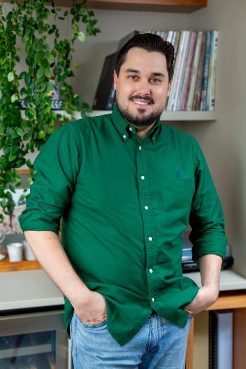 Tainan Salvador sorridente, vestindo camisa verde escura com as mãos nos bolsos da calça jeans, posando em frente a uma estante decorada. (Posicionamento de Imagem, Fotos Bruno Arita)'