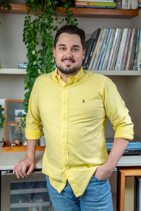 Tainan Salvador posando em pé em cenário de home bar moderno, vestindo camisa amarela clara e calça jeans. Ele está com uma mão no bolso e a outra apoiada no balcão, com plantas e discos ao fundo. (Retrato Corporativo Moderno, Fotos Bruno Arita)'
