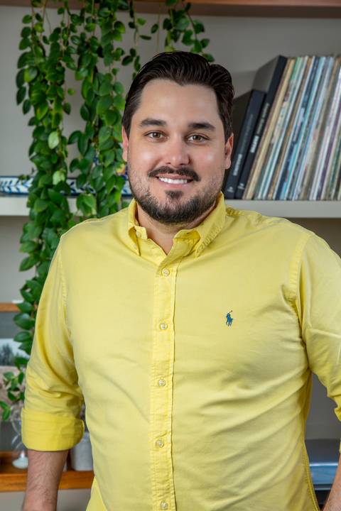 Tainan Salvador em plano médio, de braços cruzados e sorriso aberto, destacando sua personalidade leve e profissional em cenário in loco. (Retrato Corporativo, Fotos Bruno Arita)'