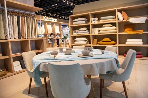 Mesa posta redonda com toalhas de mesa Karsten, xícaras e estantes de produtos ao fundo. (ABUP Show 2026, Fotos Bruno Arita)'