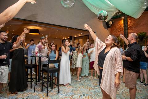 Maria e Lucas propondo um brinde com copos erguidos em plano aberto mostrando convidados ao fundo no ABC. Foto por Bruno Arita.'