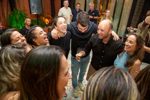 Lucas e Maria abraçados em roda de amigos em momento de celebração espontânea. Registro por Bruno Arita.'