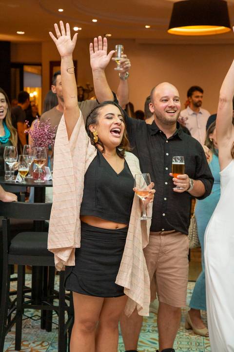Maria e Lucas dançando e comemorando com os braços para cima enquanto seguram copos. Registro de alegria por Bruno Arita.'