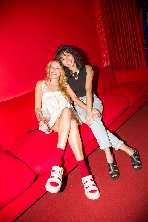 Erika e convidada posando no sofá vermelho com mood editorial de moda. Autoria: Bruno Arita.'