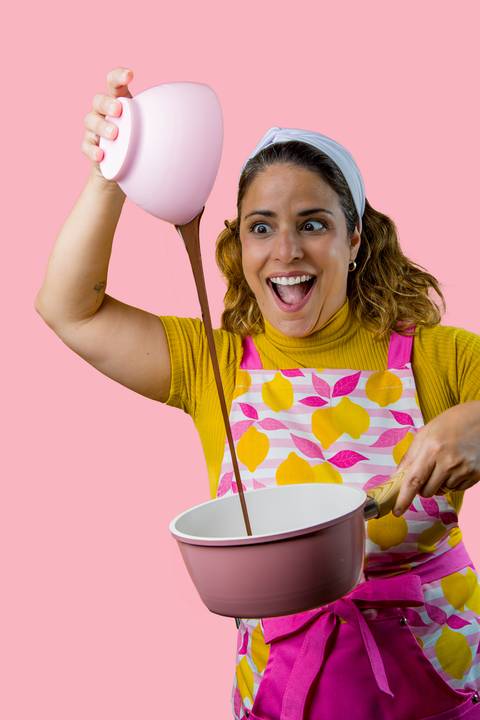Expressão de surpresa e alegria de Renata Mendes ao ver o chocolate derretido cair na panela, fundo rosa. Foto por Bruno Arita.'