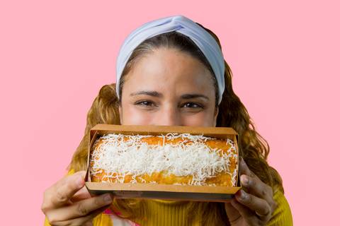 Renata Mendes sorrindo ao apresentar seu bolo de coco artesanal na caixinha, destacando o produto final. Registro por Bruno Arita.'