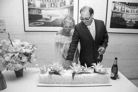 Noivos sorrindo enquanto cortam juntos a primeira fatia do bolo de casamento. Fotografia em preto e branco por Bruno Arita.'