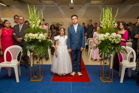 Filhos do casal posando com flores decoradas ao entrarem na cerimônia com público em volta. Autoria: Bruno Arita.'