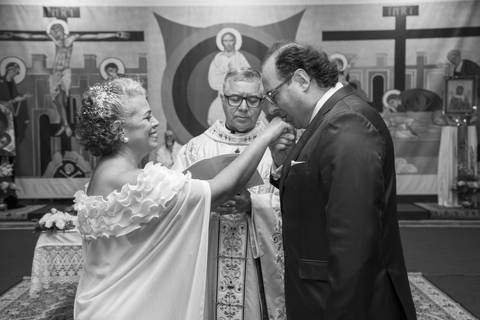 Noivo Danilo beija a mão da noiva Tabatha, que sorri espontaneamente com o padre ao fundo. Fotografia PB por Bruno Arita.'