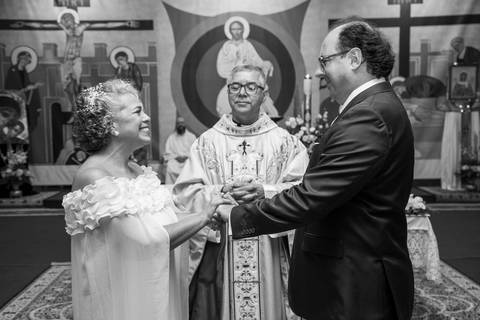 Casal sorrindo um para o outro e consagrando o casamento com o padre ao fundo. Fotografia PB por Bruno Arita.'