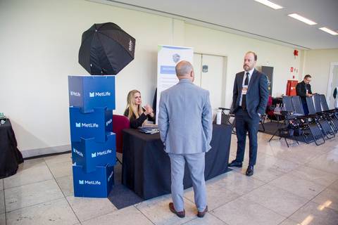 Homens de terno conversam perto de balcão com atendente loira em estande de evento. Foto institucional por Bruno Arita para SCOPO e MetLife no Centro de Convenções Rebouças.'
