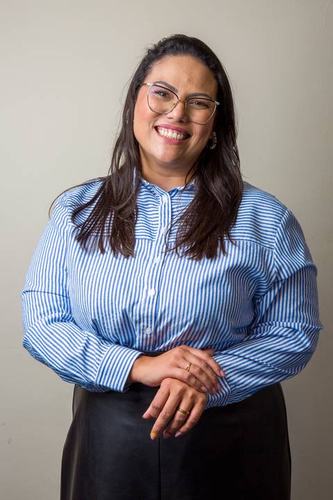 Médica sorrindo de óculos, camisa social listrada azul e branca e saia preta, com as mãos cruzadas. Foto profissional por Bruno Arita para a SCOPO MetLife no Centro de Convenções Rebouças em SP.'