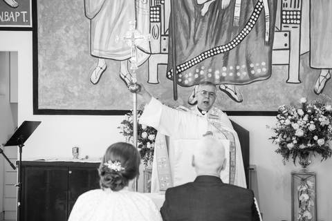 Padre abençoando o casal no altar com a mão estendida durante o sacramento do matrimônio. Foto por Bruno Arita.'