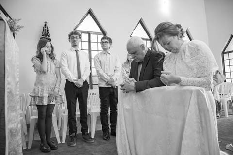 Família ajoelhada no altar recebendo a bênção especial do padre durante o casamento. Foto por Bruno Arita.'