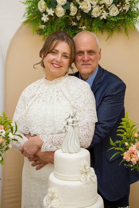 Retrato acolhedor dos noivos abraçados atrás do topo de bolo de casamento. Foto por Bruno Arita.'