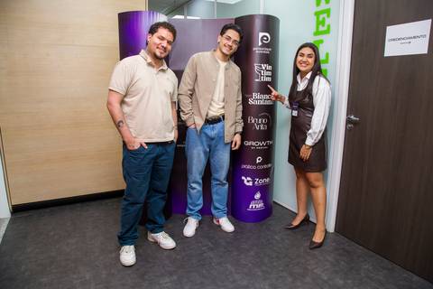 Três participantes posando felizes em frente ao painel de patrocinadores do evento. Foto por Bruno Arita.'