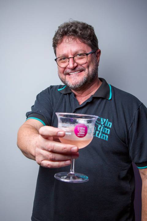 Homem sorridente vestindo camisa preta da Vintimtim fazendo um brinde com taça de drink. Foto por Bruno Arita.'