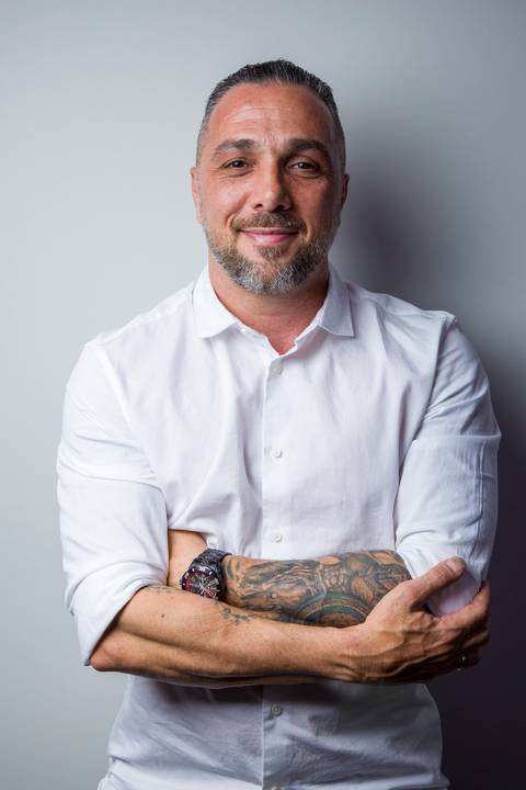 Retrato corporativo de homem de camisa branca e braços cruzados exibindo tatuagens no braço. Foto por Bruno Arita.'