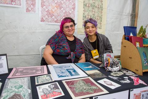 Duas artistas plásticas sentadas atrás de mesa com xilogravuras e impressões artísticas expostas. Foto por Bruno Arita.'