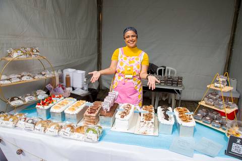 Confeiteira Renata Mendes de avental rosa posando orgulhosa atrás de mesa farta de bolos recheados e sobremesas da Bem Caseirinho SP. Foto por Bruno Arita.'