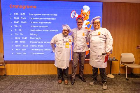 Apresentação do Cronograma: Três palestrantes caracterizados como chefs no palco à frente da tela de programação do Summit. Foto por Bruno Arita.'