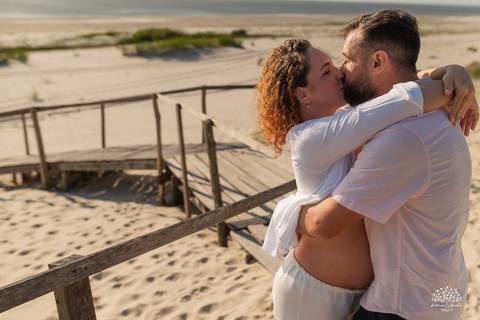 Casal se abraçando na areia da praia, com passarela ao fundo'
