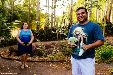 Pai vestindo camiseta do Star Wars, segurando boneco do Mestre Yoda e sapatinho branco de bebe com mãe gestante ao fundo, durante ensaio fotografico em maringa'