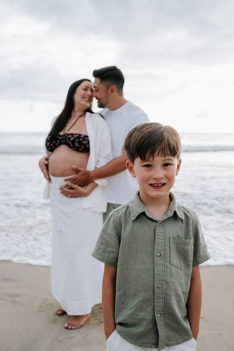 Foto do filho sorrindo em primeiro plano com os pais ao fundo na praia'