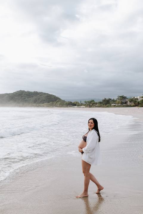 Foto da gestante em pé na beira do mar com montanhas ao fundo'