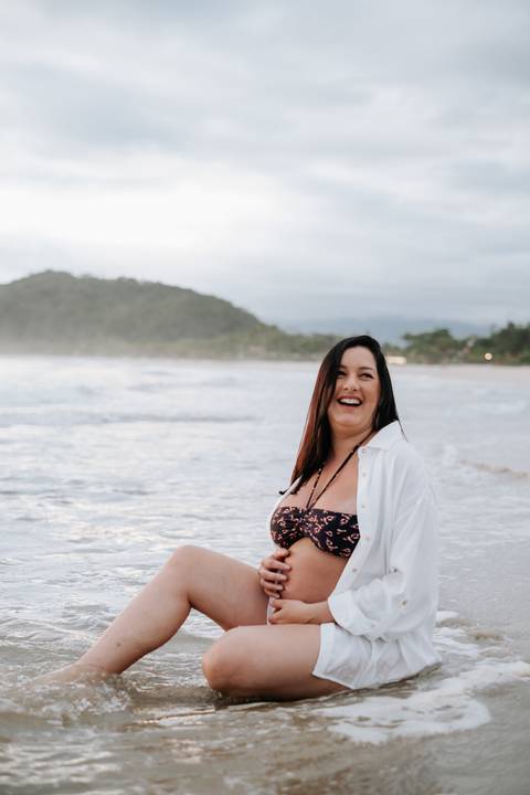 Foto da gestante sorrindo sentada na areia da praia'