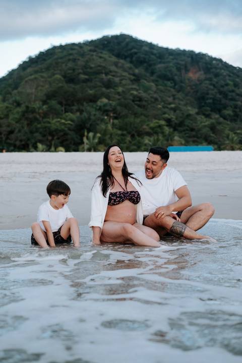 Foto da família sorrindo sentada na areia com água do mar nos pés'