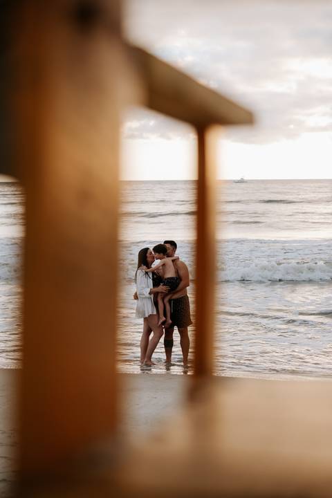 Foto da família abraçada na praia vista através de elementos de madeira'