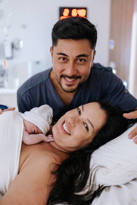 Foto do casal sorrindo com o bebê recém-nascido no colo.'