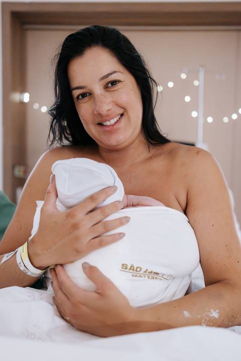 Foto da mãe segurando o recém-nascido enrolado em manta branca.'