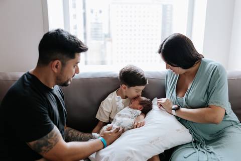 Foto dos pais e o filho mais velho com o recém-nascido no sofá.'