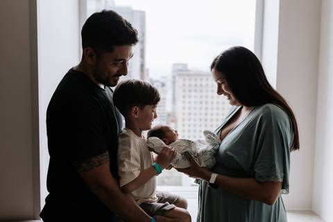 Foto da família sorrindo com o recém-nascido na janela da maternidade.'