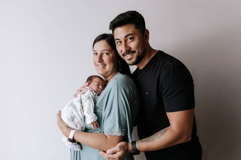 Foto dos pais sorrindo com o bebê no quarto do hospital.'