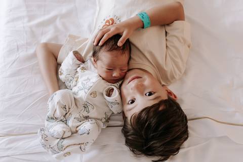 Foto do irmão mais velho deitado na cama ao lado do bebê.'