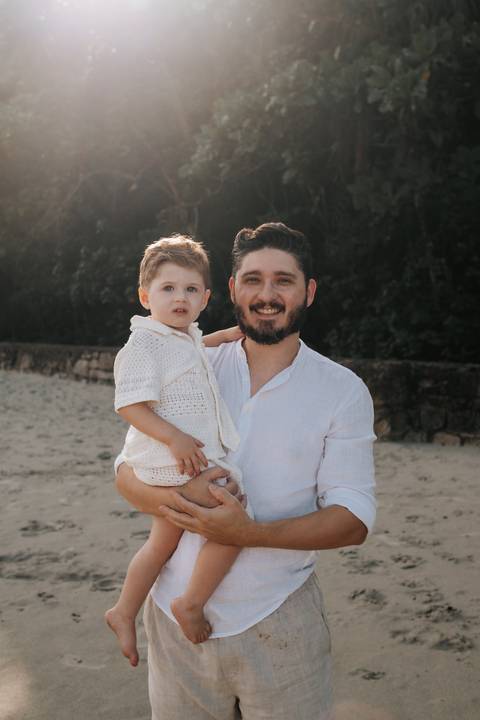 Foto do pai com o filho no colo em ensaio gestante na praia.'